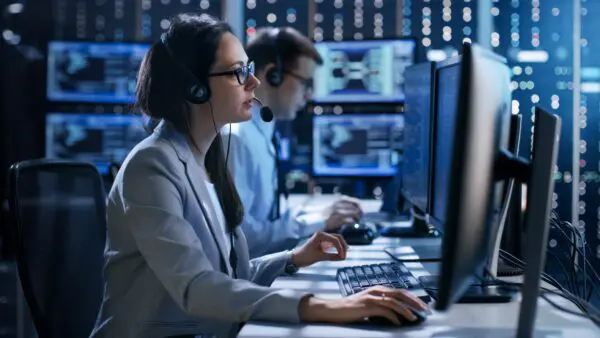 Female working in a Technical Support Team Gives Instructions with the Help of the Headsets. In the Background People Working and Monitors Show Various Information.