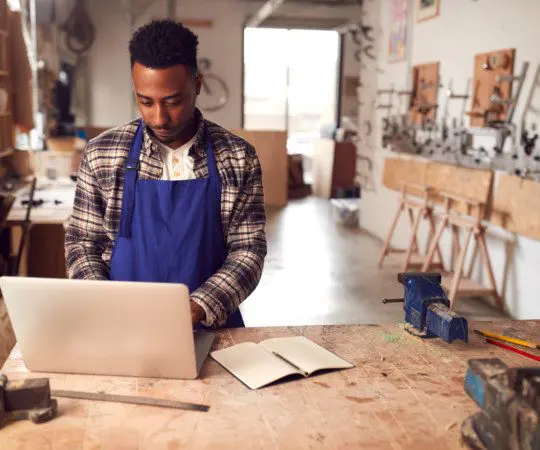 Male Craftsman In Carpentry Workshop For Bamboo Bicycles  Doing Accounts On Laptop